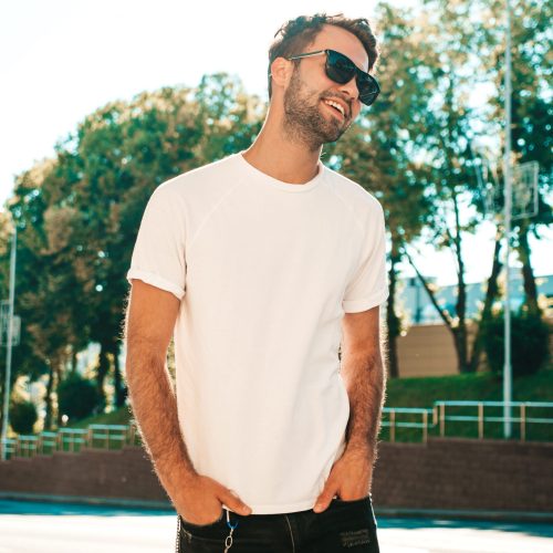 Portrait of handsome smiling stylish hipster lambersexual model.Man dressed in white T-shirt. Fashion male posing on the street background in sunglasses outdoors